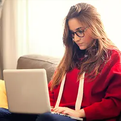 		 Foto em ambiente interno. Mulher jovem, com cabelos lisos, usando óculos, blusa de frio vermelha e calça escura, sentada no sofá com um notebook em seu colo. Ela está olhando para a tela do notebook.