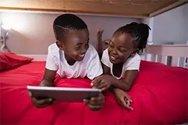 Imagem de duas crianças deitadas em uma cama com um tablet em mãos sorrindo uma para a outra.