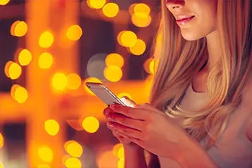 Moça com cabelos loiros olhando para um celular. Na imagem, aparece somente a boca e o nariz. Tem muitas luzes no fundo.