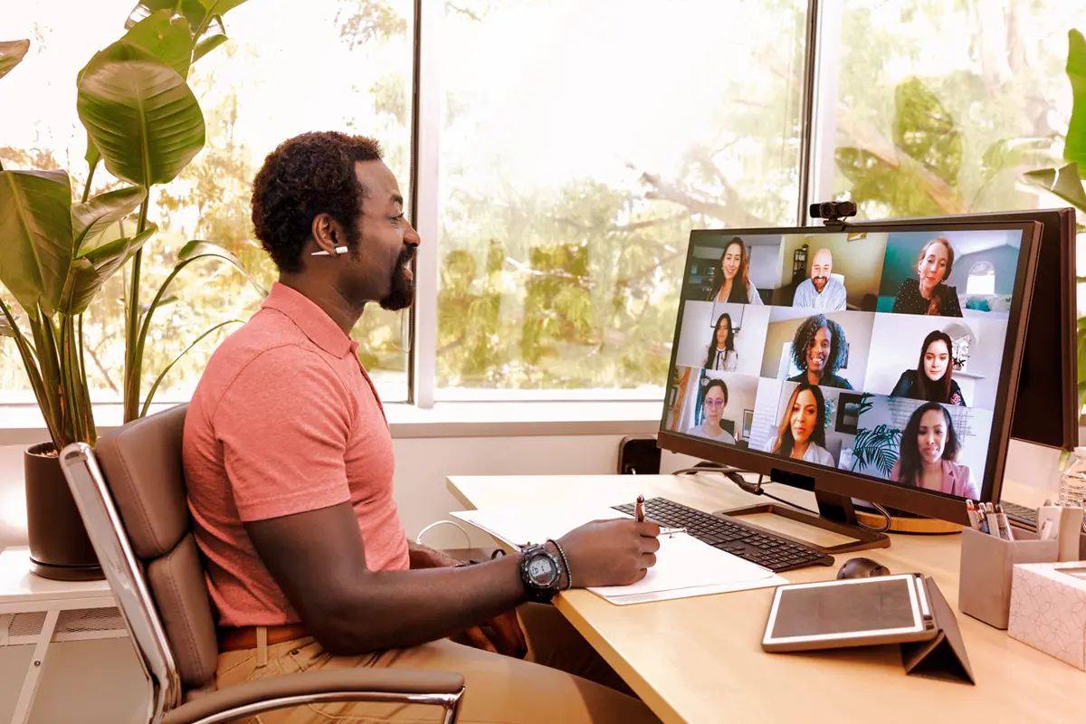 Homem com fones de ouvido em uma videochamada de trabalho, sentado em sua mesa de home office com um computador exibindo uma reunião virtual com várias pessoas. O uso de uma conexão de internet corporativa permite a realização de chamadas de vídeo estáveis.