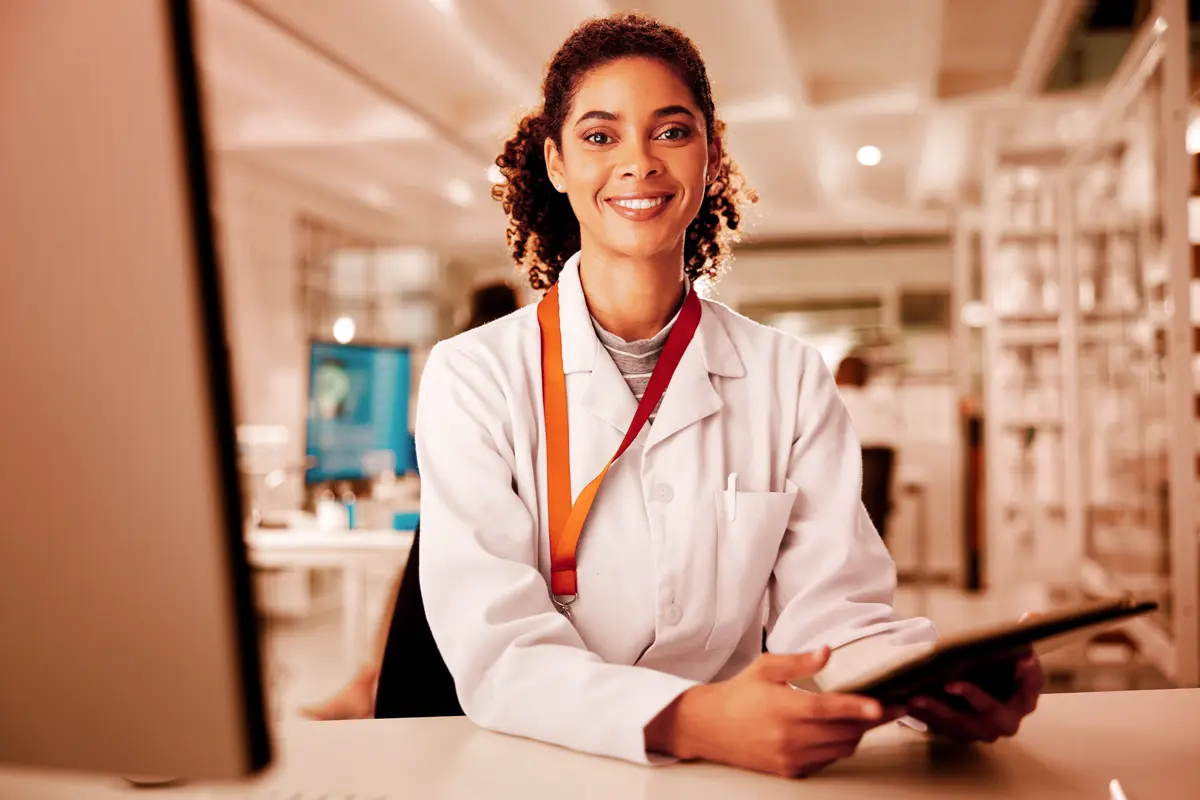Uma profissional de saúde sorrindo, vestindo jaleco e segurando um tablet, está em sua mesa com um computador, em um ambiente de laboratório ou consultório. A imagem ilustra o conceito de telemedicina corporativa em um ambiente de trabalho profissional, onde a tecnologia é utilizada para cuidados de saúde e consultas remotas.