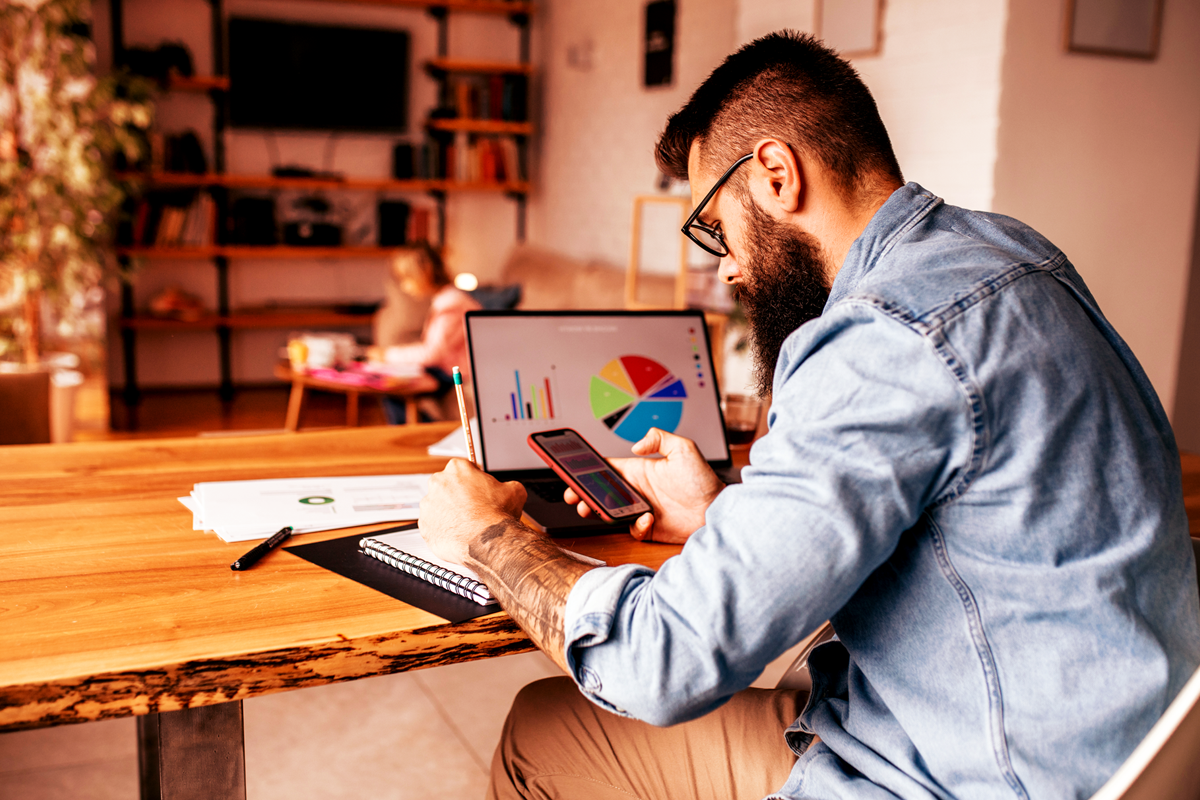Homem barbudo com camisa jeans trabalha em home office sobre uma mesa de madeira, conferindo o celular enquanto anota em um caderno; ao fundo, um notebook exibe gráficos de barras e um gráfico de pizza colorido, ilustrando organização e produtividade com Google Workspace para empresas em um ambiente quente e aconchegante, com sala e estante desfocadas ao fundo.
