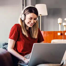 Foto em ambiente interno. Mulher jovem, com cabelos lisos, usando óculos, blusa vermelha e fones de ouvido, sentada no sofá com um notebook em seu colo. Ela está olhando para a tela do notebook.