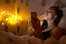 Imagem de uma moça sentada em uma cama com um celular e fones de ouvido. O ambiente está com pouca luz.