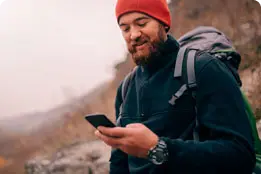 Homem jovem, de barba e roupas de inverno, está em uma montanha, olhando o celular.
