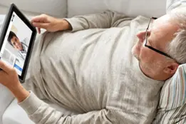 Imagem de um senhor de cabelos brancas deitado no sofá com um ipad em mãos.