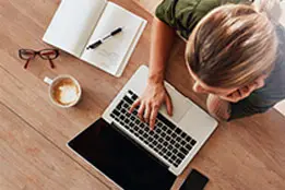 Foto tirada pelo ângulo de cima de uma mesa onde tem uma mulher usando notebook. Ao lado tem uma xícara de café, um óculos, uma caderno de anotações, uma caneta e um celular.