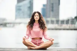 Foto em ambiente externo com mulher jovem de olhos fechados e sentada em posição de meditação. Ela tem pele clara, cabelos escuros, longos e ondulados, veste blusa de manga longa e shorts. Ao fundo se vê a paisagem de uma cidade grande com prédios em plano desfocado.