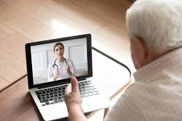Um senhor de cabelos brancos com um laptop na frente dele com a imagem de uma médica na tela.