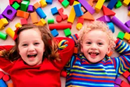 Foto de duas crianças deitadas no chão em cima de pecinhas coloridas de brinquedo. Elas estão rindo.