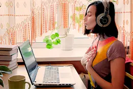 Imagem de uma moça com fones de ouvido em frente a um laptop. Ela esta de olhos fechados e mãos no peito.