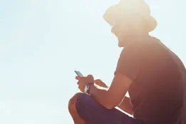 Imagem de um homem com chapéu e óculos escuros, sorrindo, sentado apoiando o braço na perna e mexendo no celular, no fundo céu azul e raios de sol refletindo na cabeça do homem.
