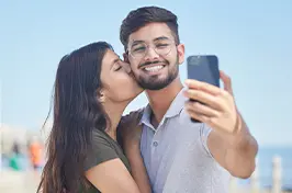 Imagem de uma moça beijando o rosto do rapaz enquanto ele usa o celular para tirar fotos.