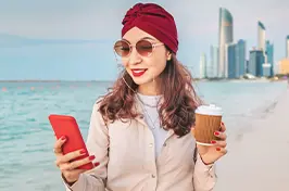 Imagem de uma mulher usando o celular com uma praia e grandes prédios no fundo.