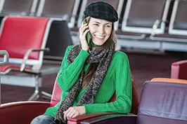 Imagem de um moça usando boina sentada em bancos de aeroporto falando ao celular.