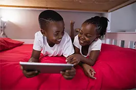 Imagem de duas crianças deitadas em uma cama com um tablet em mãos sorrindo uma para a outra.