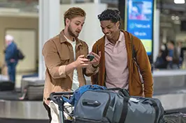 Dois rapazes jovens conversando com malas em um ambiente de aeroporto enquanto consultam informações em um smartphone.