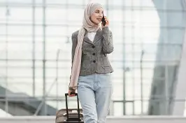Imagem de uma moça sorrindo falando ao celular, lenço na cabeça e uma mala.
