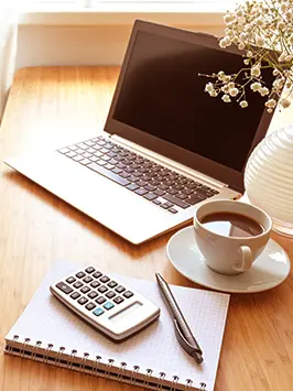 Imagem de um computador sob a mesa com um caderno, uma calculadora e uma xícara de café.