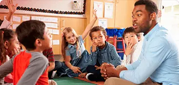 Um adulto sentado em circulo tirando duvida de varias crianças com as mãos levantadas.
