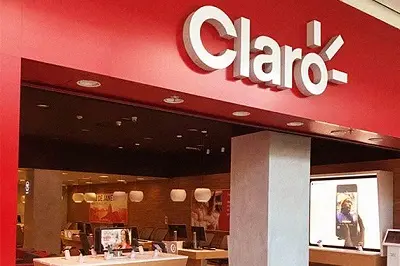 Fachada de uma loja Claro. Parede vermelha com o logo da Claro em branco. Mesa com exposição de celulares no interior da loja.
