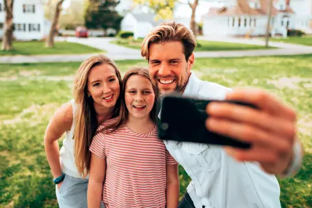 Imagem de uma família de cabelos claros sorrindo e fazendo uma foto com um smartphone em um gramado.