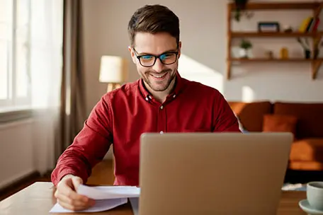 Homem sorridente olhando para uma tela de notebook com algumas folhas de papel na mão.