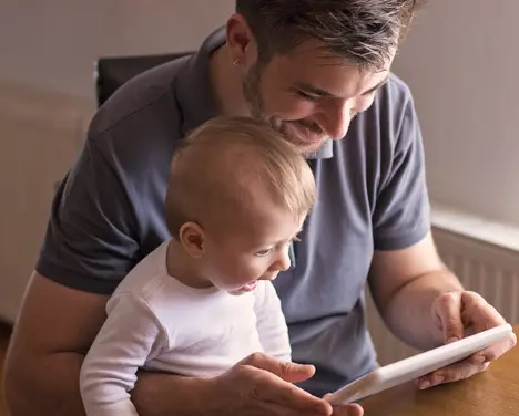 Imagem de um pai com um bebê no colo mexendo em um tablet.