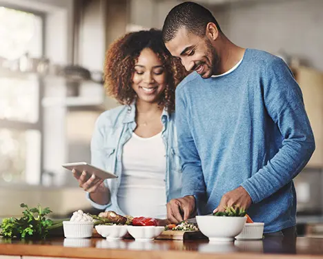 Imagem de um casal cozinhando juntos. a Moça segura um celular na mão enquanto o homem prepara os alimentos.