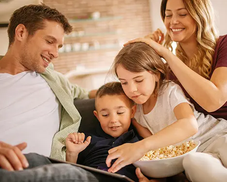 Imagem de uma família com marido, esposa e dois filhos comendo pipoca e mexendo num tablet.