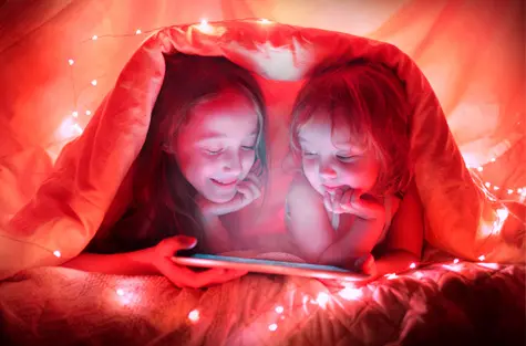 Imagem de duas crianças embaixo de uma coberta com um tablet nas mãos. O ambiente tem muitas luzes.