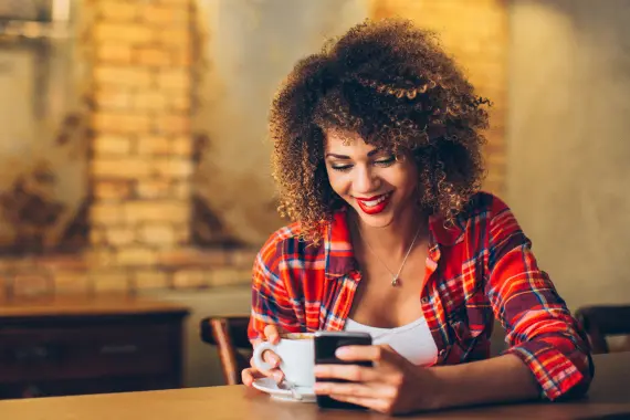 Moça sorrindo de cabelos cacheados e camisa xadrez com uma xícara de café em uma mão e segurando um celular na outra.