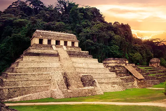 imagem: pirâmides do México na frente e árvores com a copa verde atrás e alguns raios solares no céu.